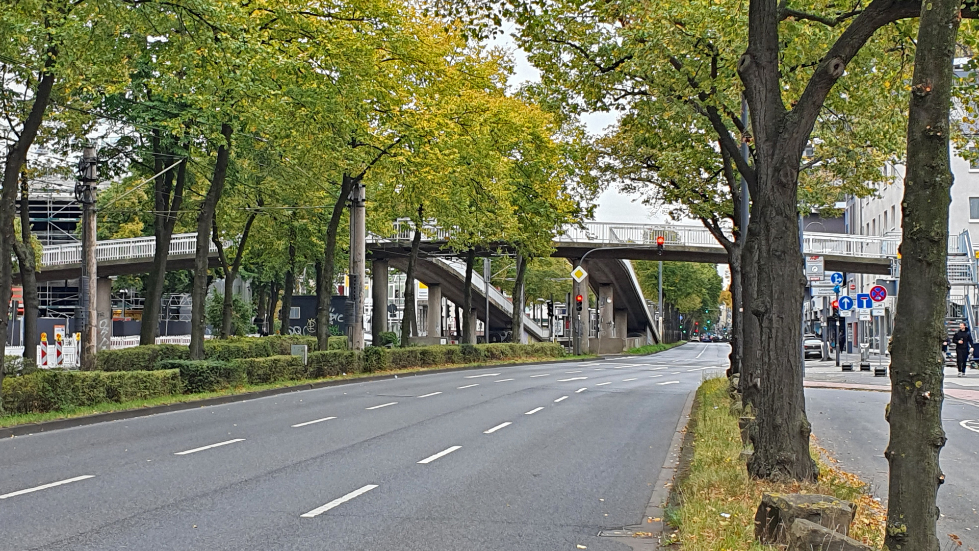 Fußgängerbrücke über die Luxemburger Straße in Köln, Blick stadtauswärts. Die Brücke verläuft schräg über die Straße, ist von Bäumen gesäumt und führt auf beiden Seiten über lange Rampen hinauf. Die Straße darunter ist mehrspurig und wirkt leer. Im Hintergrund sind Baustellenabsperrungen zu sehen.