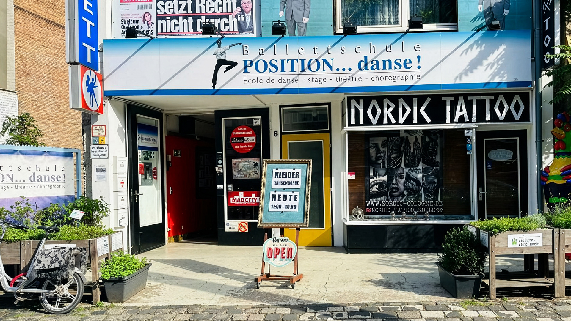 Eingangsbereich des Nachbarschaftsvereins schmitzundkunst e.V. in Köln. Das Gebäude beherbergt mehrere Einrichtungen: links die Ballettschule „POSITION… danse!“ mit blauer Beschilderung, daneben das Tattoo-Studio „Nordic Tattoo“ mit markantem schwarzen Schriftzug. Vor dem Eingang steht ein Schild mit der Aufschrift „Kleider-Tauschbörse – Heute 12:00–18:00“. Pflanzenkübel und ein Lastenfahrrad schmücken den Eingangsbereich.