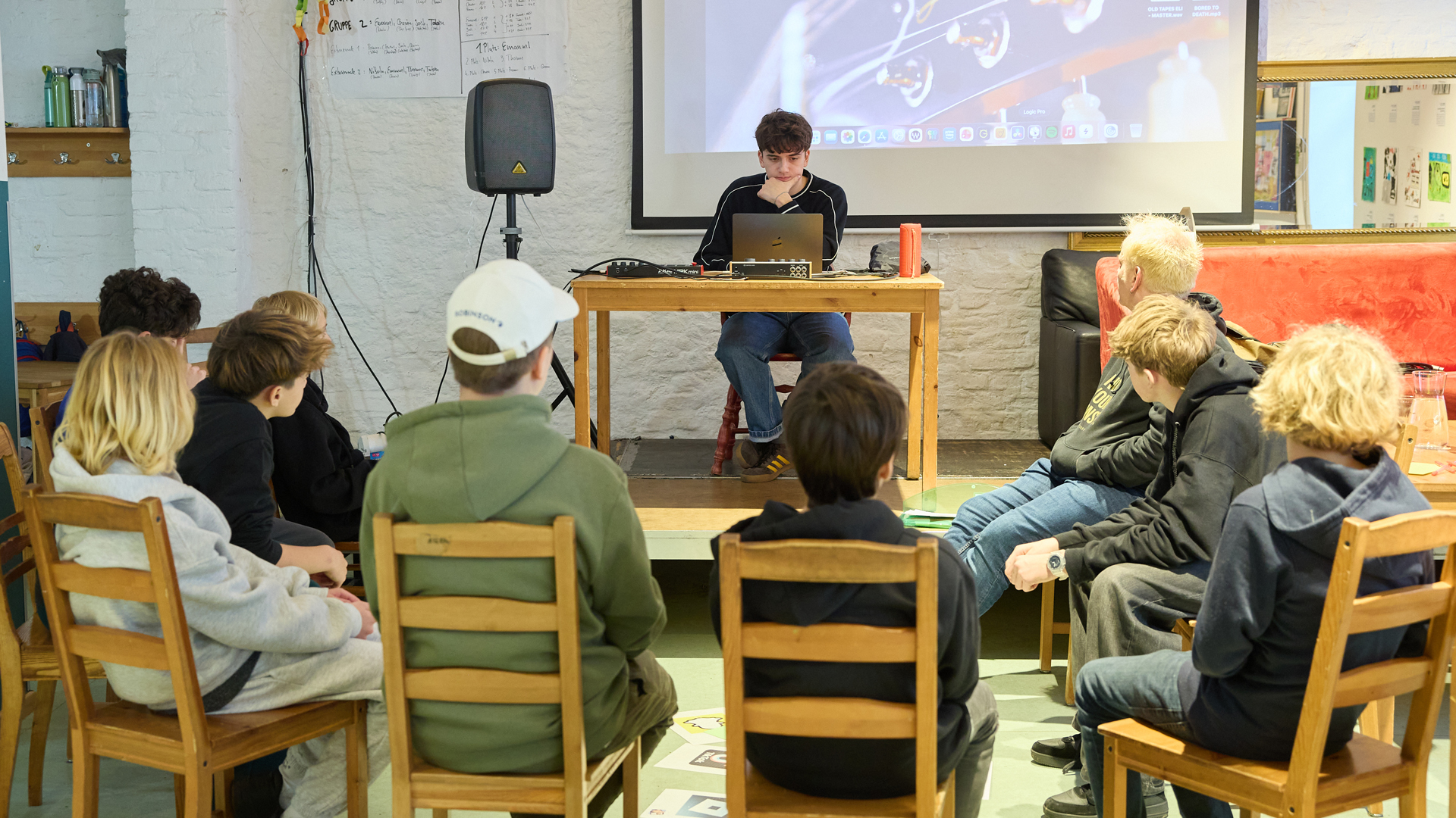 Mehrere Jugendliche sitzen im Halbkreis auf Holzstühlen und hören aufmerksam einem jungen Musikstudenten zu, der vorne an einem Tisch mit Laptop und Mischpult sitzt. Im Hintergrund ist eine Leinwand mit einer Projektion und eine weiße Wand mit Plakaten zu sehen.