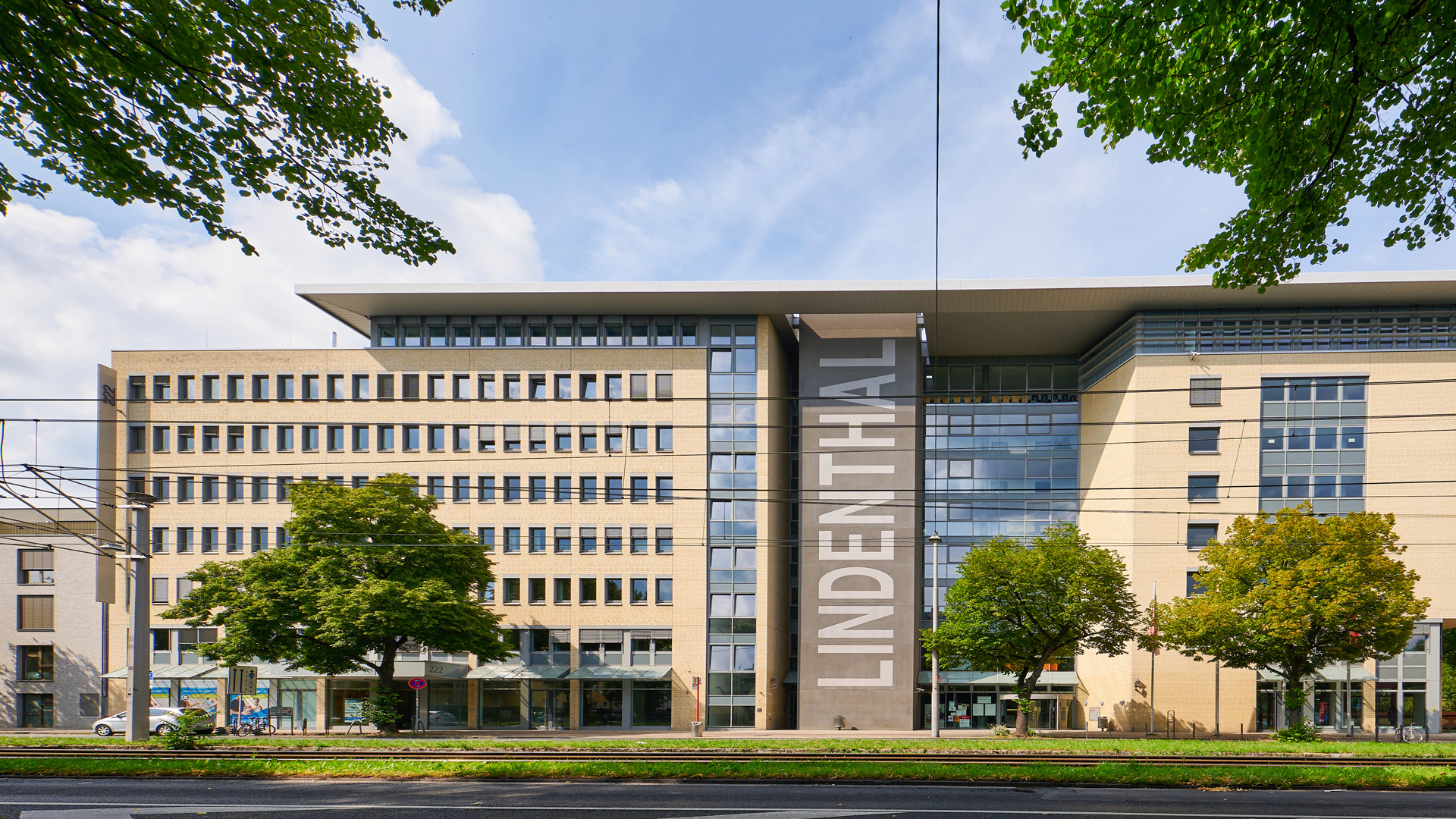 Außenansicht des Bezirksrathauses Lindenthal in Köln. Das moderne Gebäude erstreckt sich über mehrere Etagen und hat eine helle Fassade mit großen Fenstern. In der Mitte prangt gut sichtbar der Schriftzug „LINDENTHAL“ in weißen Großbuchstaben auf einer grauen Wandfläche. Vor dem Gebäude stehen grüne Bäume, im Vordergrund verlaufen Straßenbahnschienen und eine Straße. Der Himmel ist leicht bewölkt.