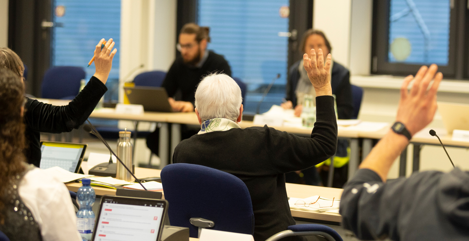 Mehrere Personen sitzen in einem Sitzungssaal an Tischen und heben die Hand zur Abstimmung, während im Hintergrund zwei Personen an einem Tisch die Sitzung leiten; auf den Tischen liegen Laptops, Unterlagen und Mikrofone.