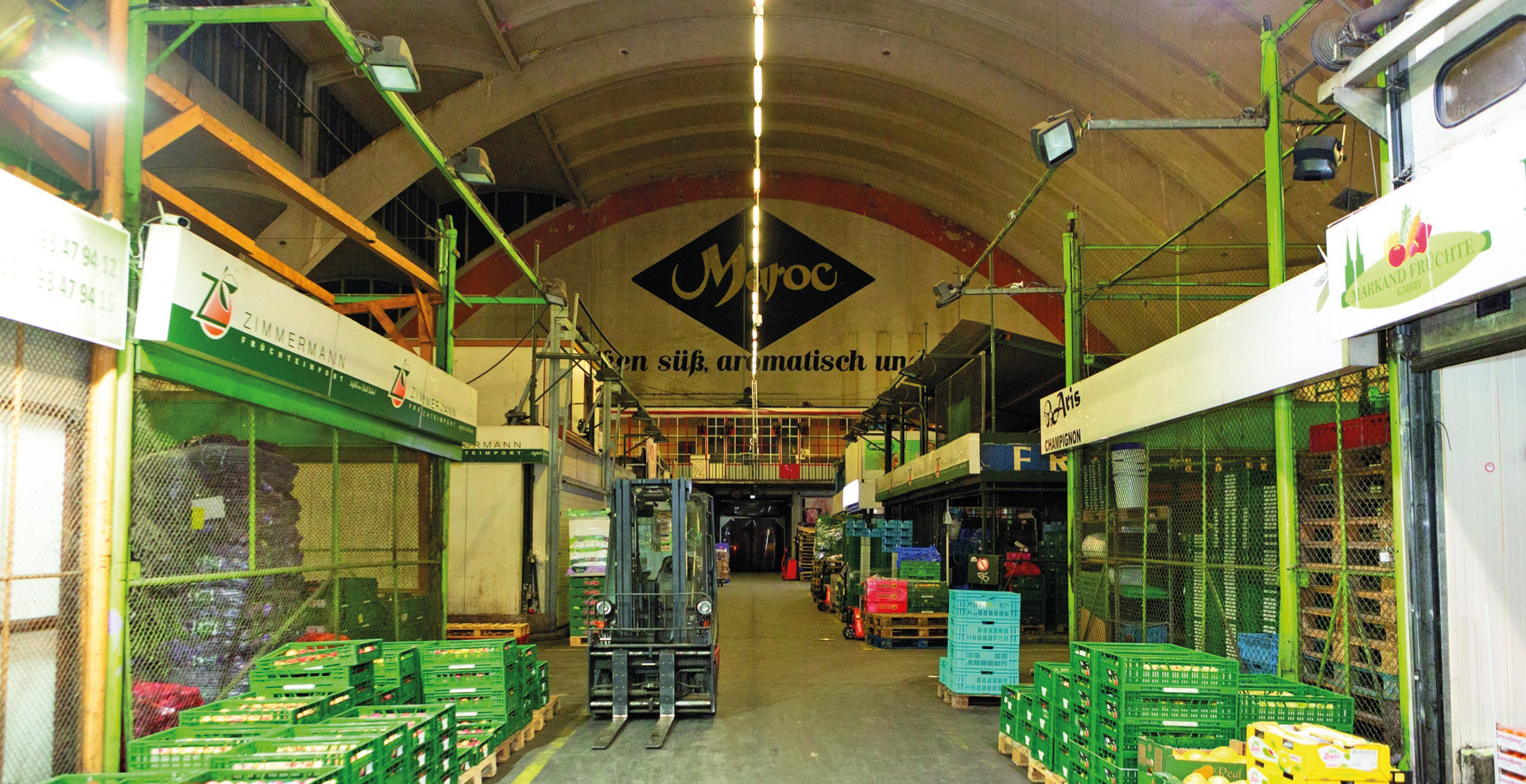 Großmarkthalle mit hohen Gewölbedecken, Paletten voller Obst- und Gemüsekisten sowie einem Gabelstapler im Mittelgang.