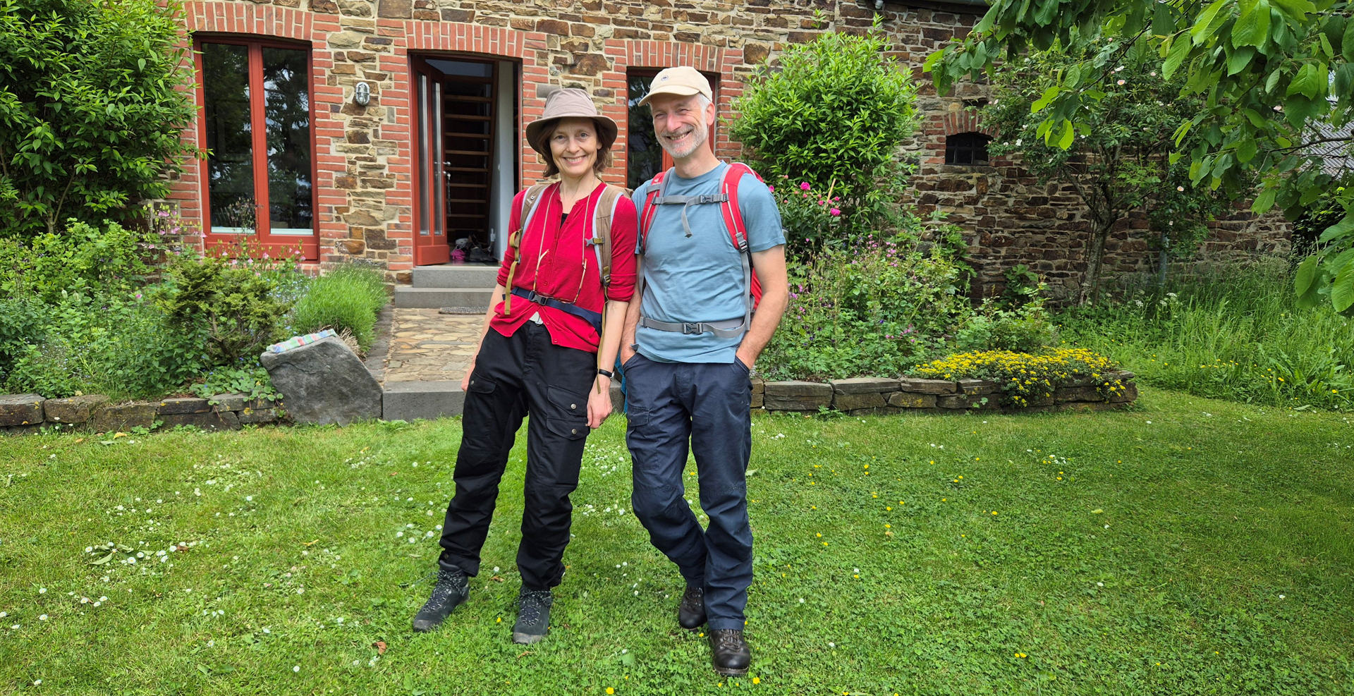 Paar mit Wanderausrüstung vor einem liebevoll renovierten Natursteinhaus im Grünen