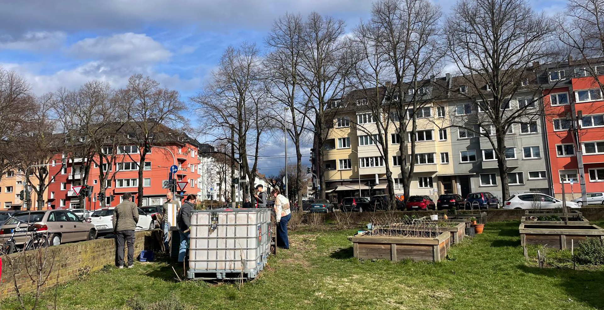 Mehrere Aktive versammeln sich im Urban-Gardening-Bereich des Sülzer Triangels rund um große Wassertanks und Hochbeete. Im Hintergrund sind Wohnhäuser und eine Straßenecke mit Bäumen zu sehen.