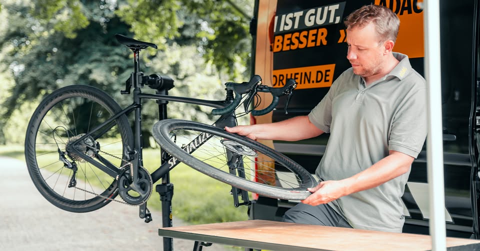 Ein Mann überprüft das Vorderrad eines Rennrads, das an einem mobilen Fahrradständer befestigt ist. Im Hintergrund ist ein Fahrzeug mit ADAC-Beschriftung zu sehen, die Szene spielt im Freien zwischen Bäumen.