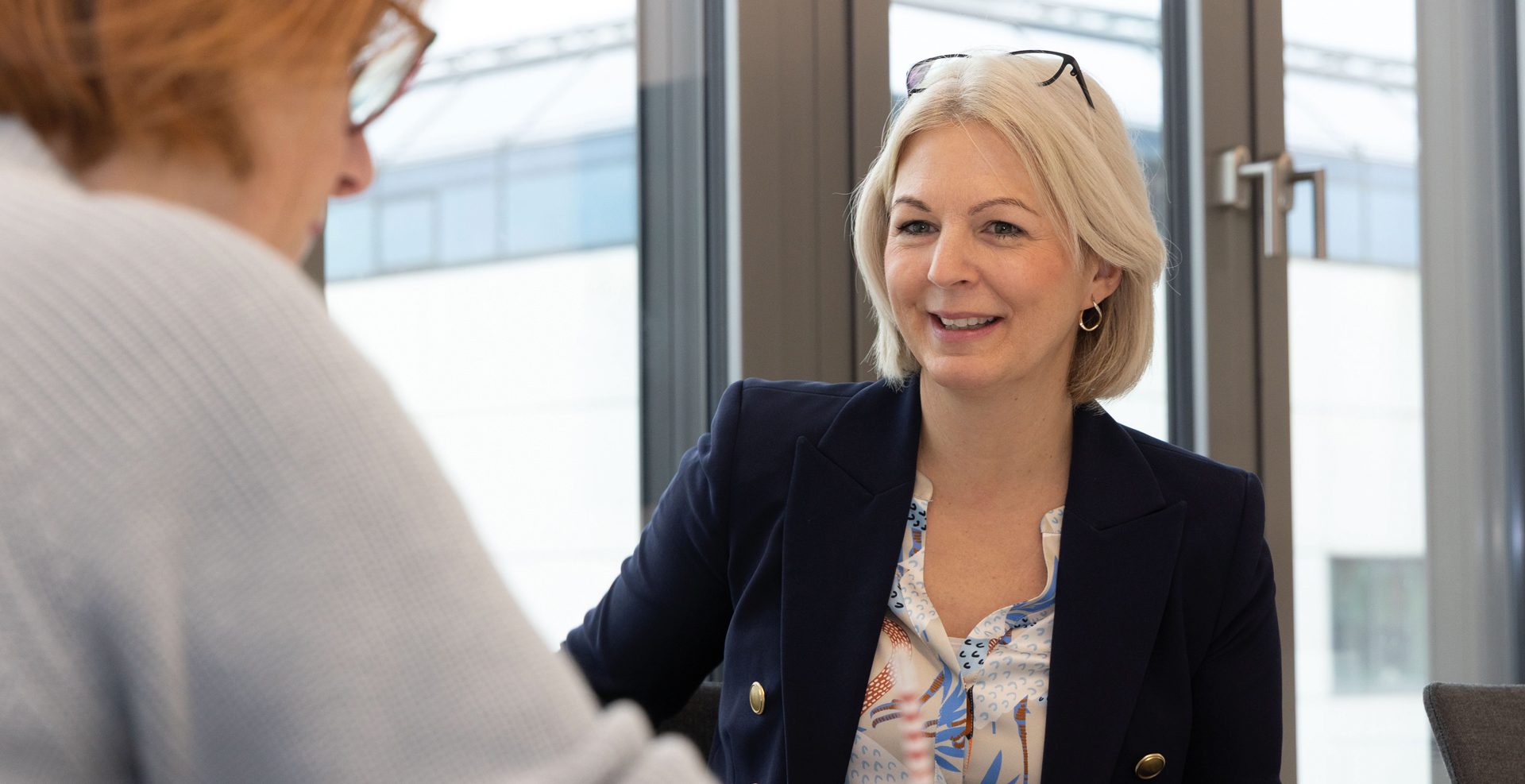Eine Frau im Business-Outfit im Gespräch mit einer anderen Person an einem Tisch vor großen Fenstern, in freundlicher, offener Atmosphäre eines Büros.