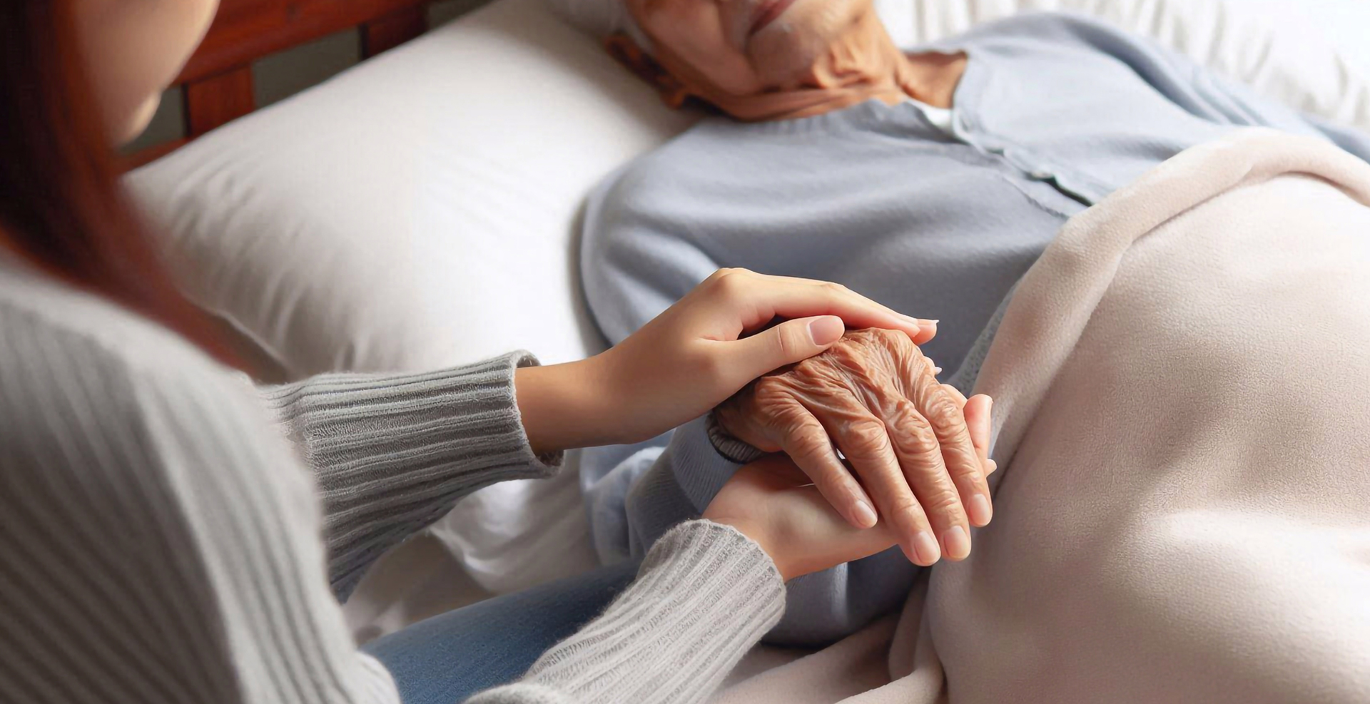 Zärtliche Geste der Fürsorge: Eine Person hält behutsam die Hand einer älteren, im Bett liegenden Frau – ein stiller Moment voller Nähe und Unterstützung.