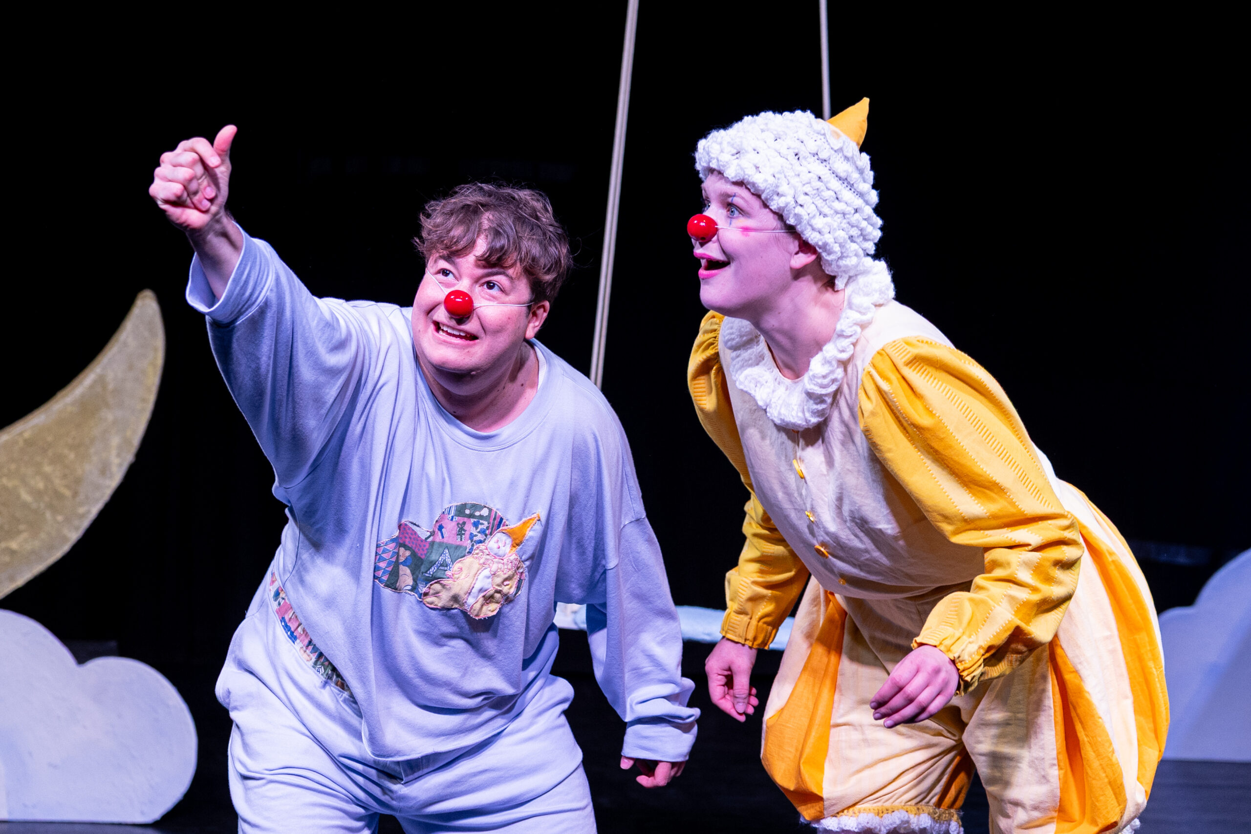 Zwei Schauspieler*innen mit roten Clownsnasen stehen nah beieinander auf der Bühne; eine zeigt begeistert nach oben, während die andere gespannt in dieselbe Richtung blickt.