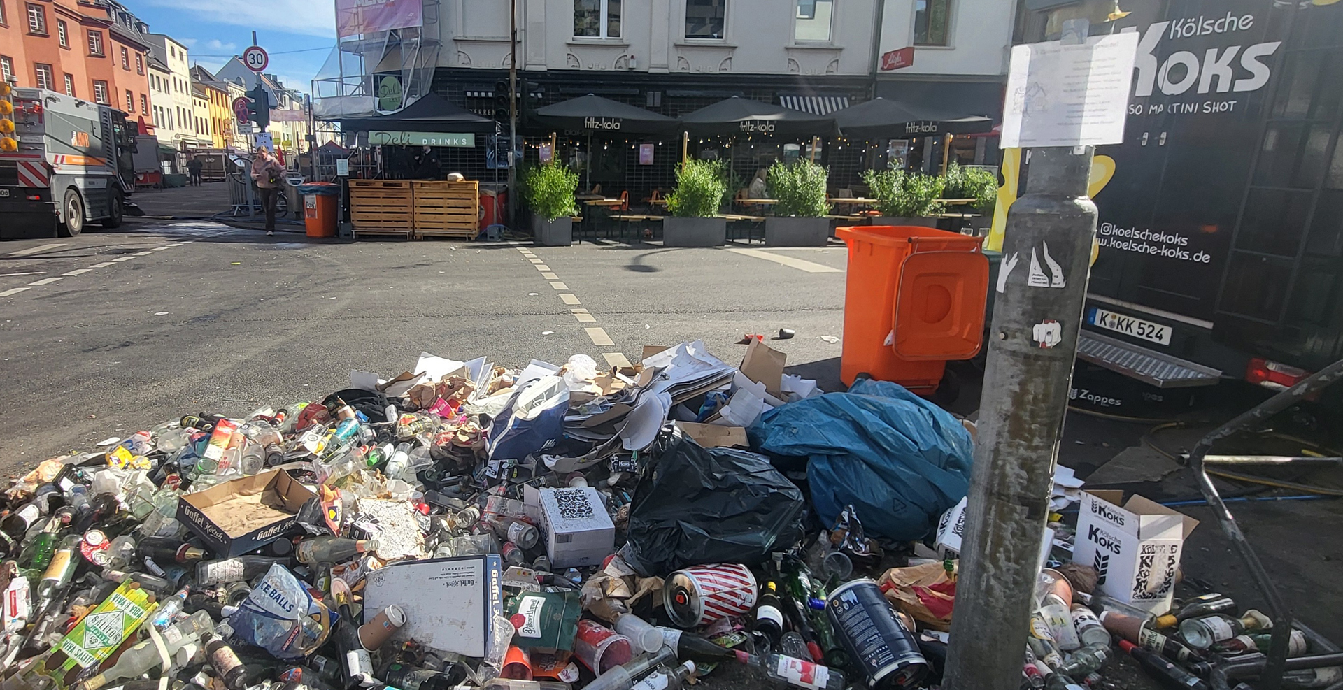 Müllberge aus Glasflaschen, Dosen, Verpackungen und Abfallsäcken liegen nach einer Veranstaltung auf einer Kreuzung an der Berrenrather Straße in Köln-Sülz. Im Hintergrund sind Gastronomiebetriebe, Müllcontainer und Einsatzfahrzeuge zu sehen.