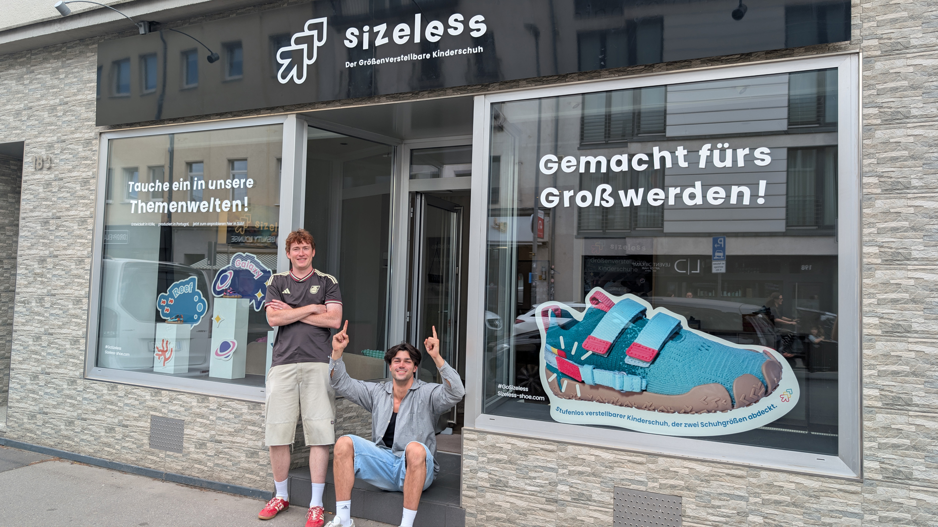 Zwei junge Gründer stehen und sitzen lächelnd vor dem Pop-up-Store von Sizeless auf der Berrenrather Straße in Köln-Sülz. Über dem Eingang ist das Logo des Startups zu sehen, in den Schaufenstern werben große Schriftzüge und eine überdimensionale Abbildung eines türkisfarbenen Kinderschuhs für den größenverstellbaren Schuh. Die beiden zeigen mit ausgestreckten Armen auf das Geschäft und vermitteln die Freude über die Eröffnung des Stores.